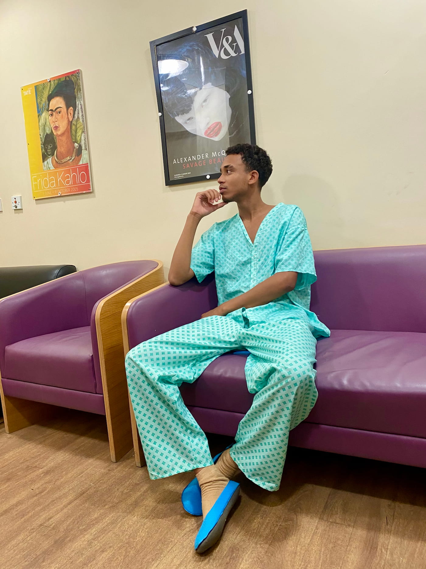 Joshua Arthur JJDREAMSS 23 years old seated in hospital waiting room Newham Centre for Mental Health London July 21 2022 wearing NHS hospital gown thoughtful Frida Kahlo Alexander McQueen Savage Beauty posters